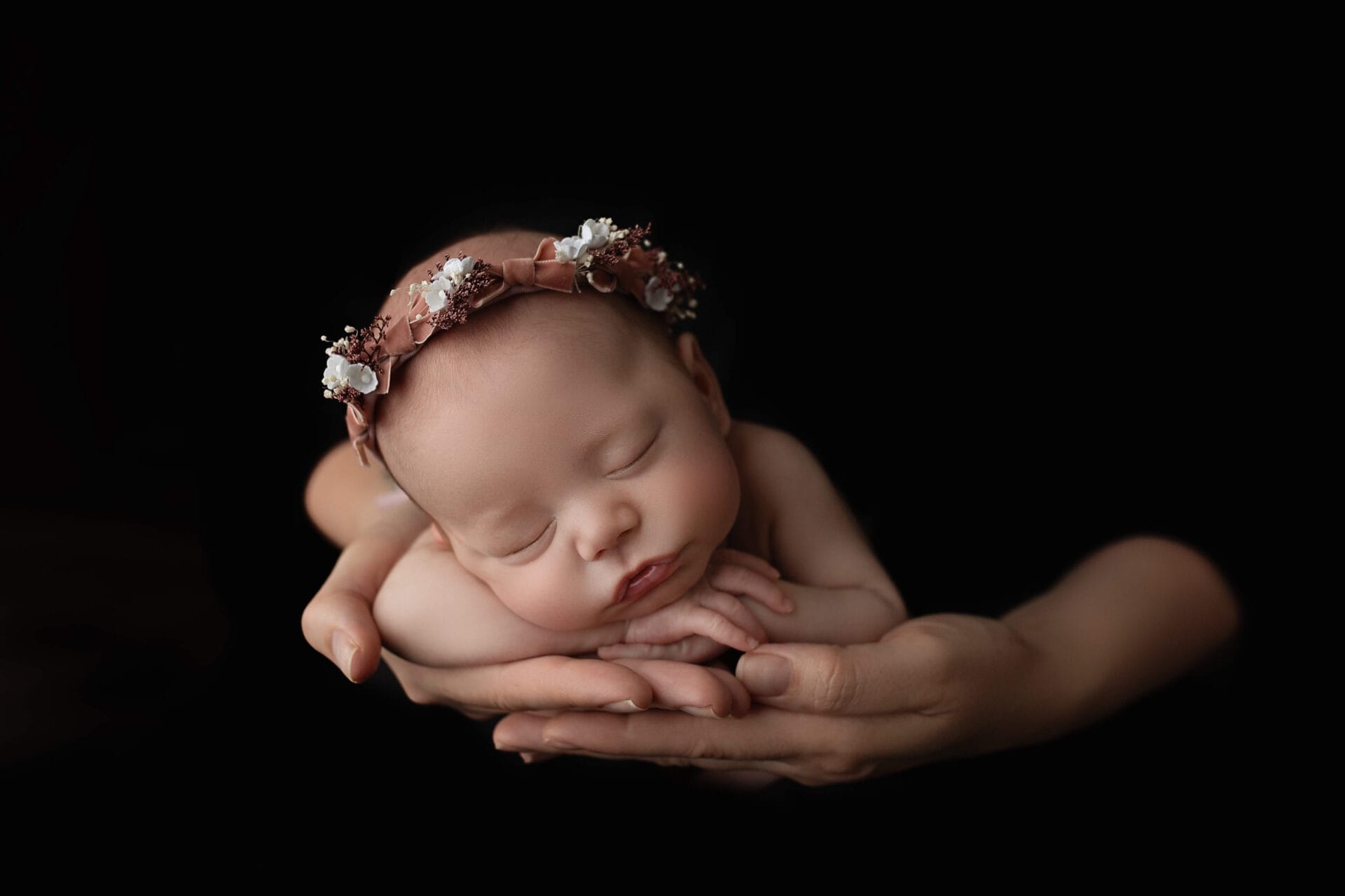 A newborn baby girl posed in her mom's hand on a black backdrop, wearing a pink floral headband.