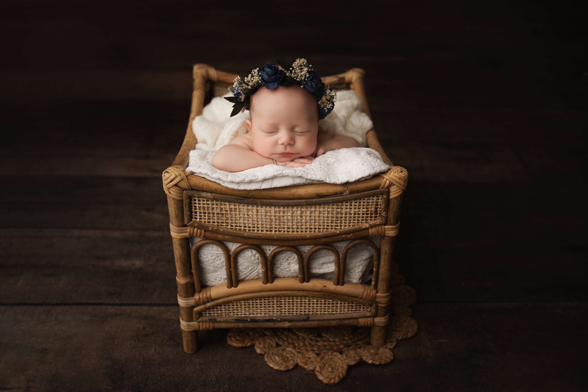 A newborn baby girl wearing a blue floral headband posed with her hands under her chin in a bamboo prop.