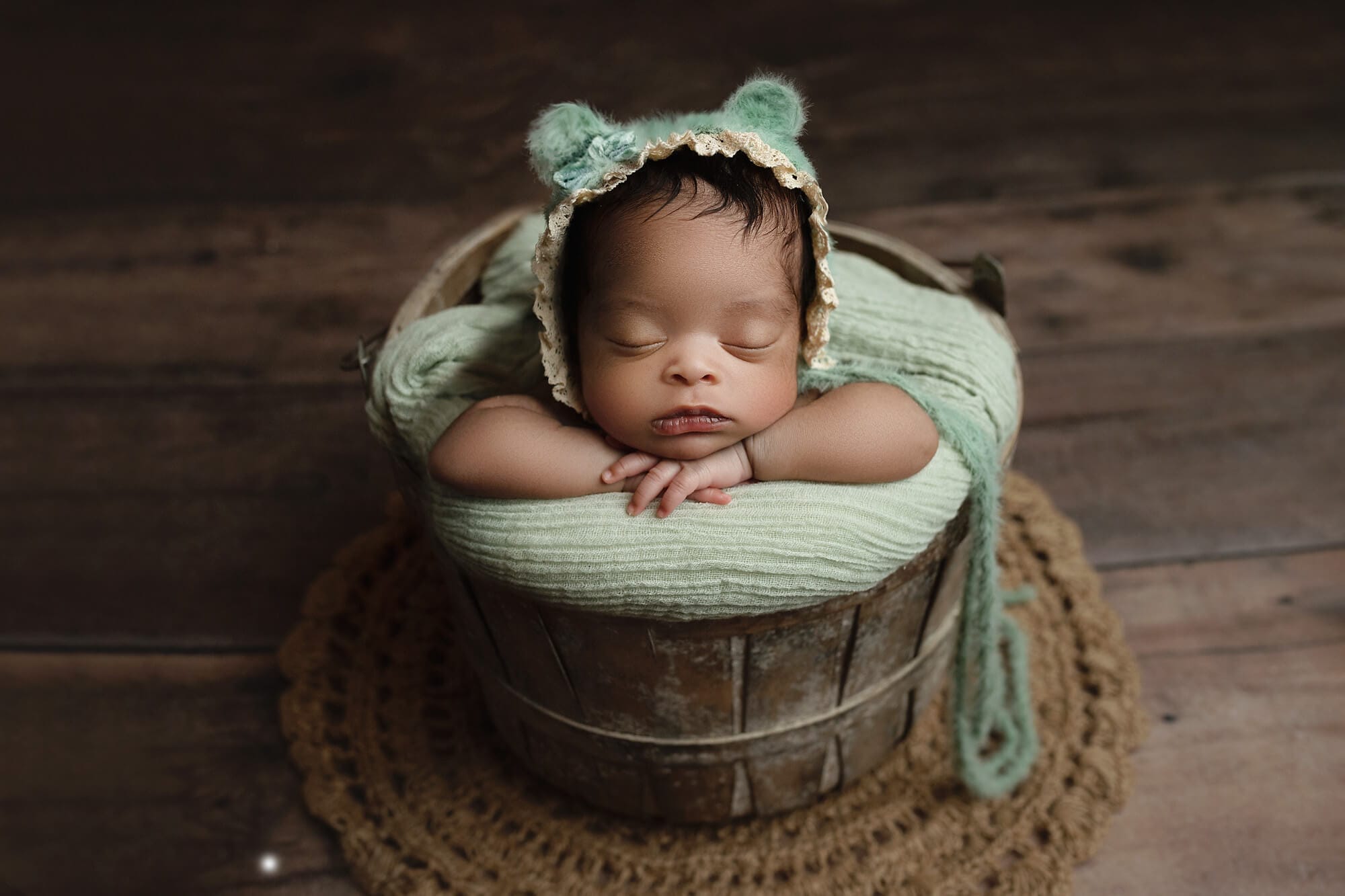 A newborn baby girl in a wooden bucket wearing a mint green bear bonnet.