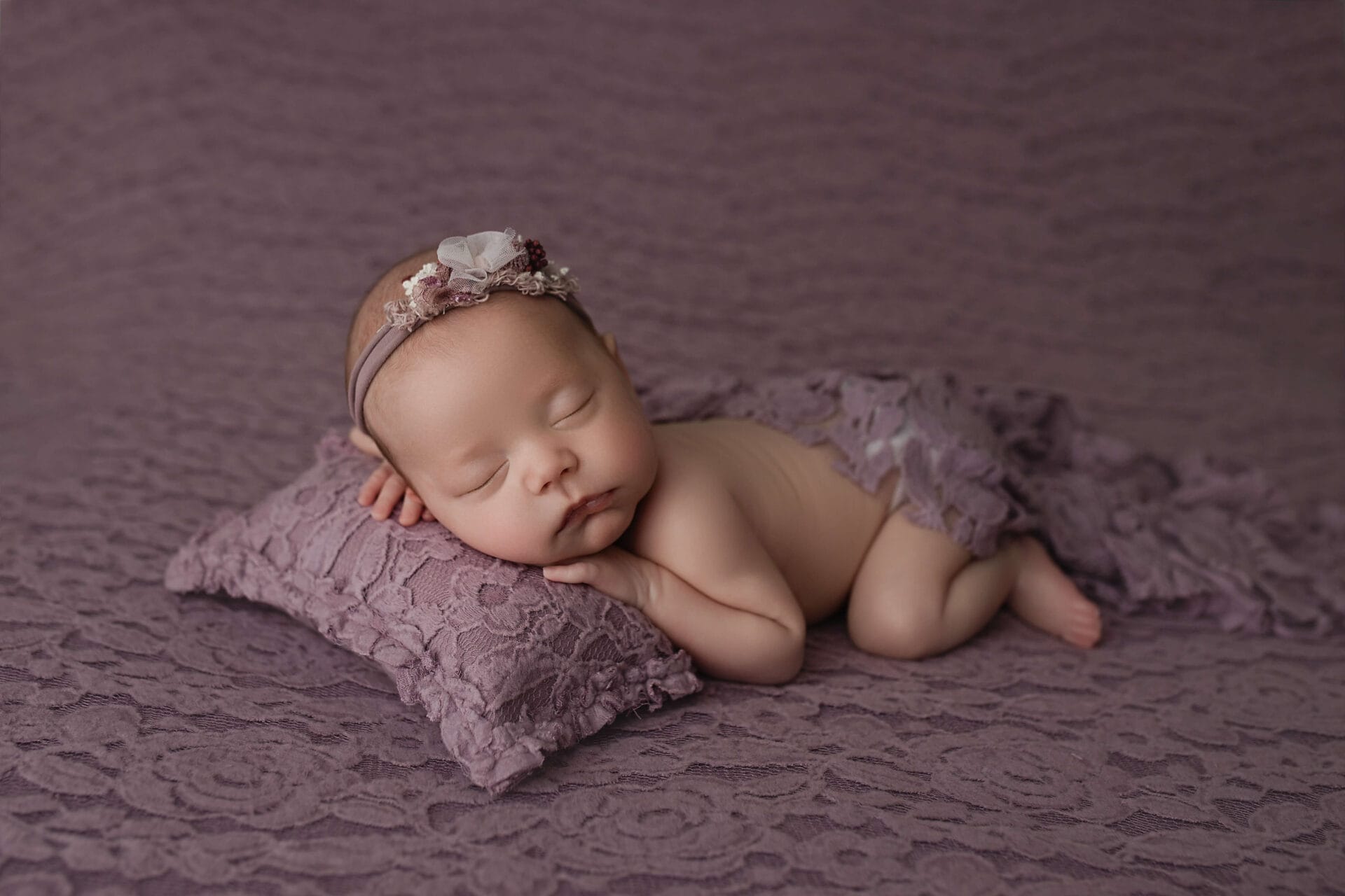 In the studio, a newborn baby girl lying on her tummy on a purple lace backdrop posed on a pillow wearing a purple floral headband.