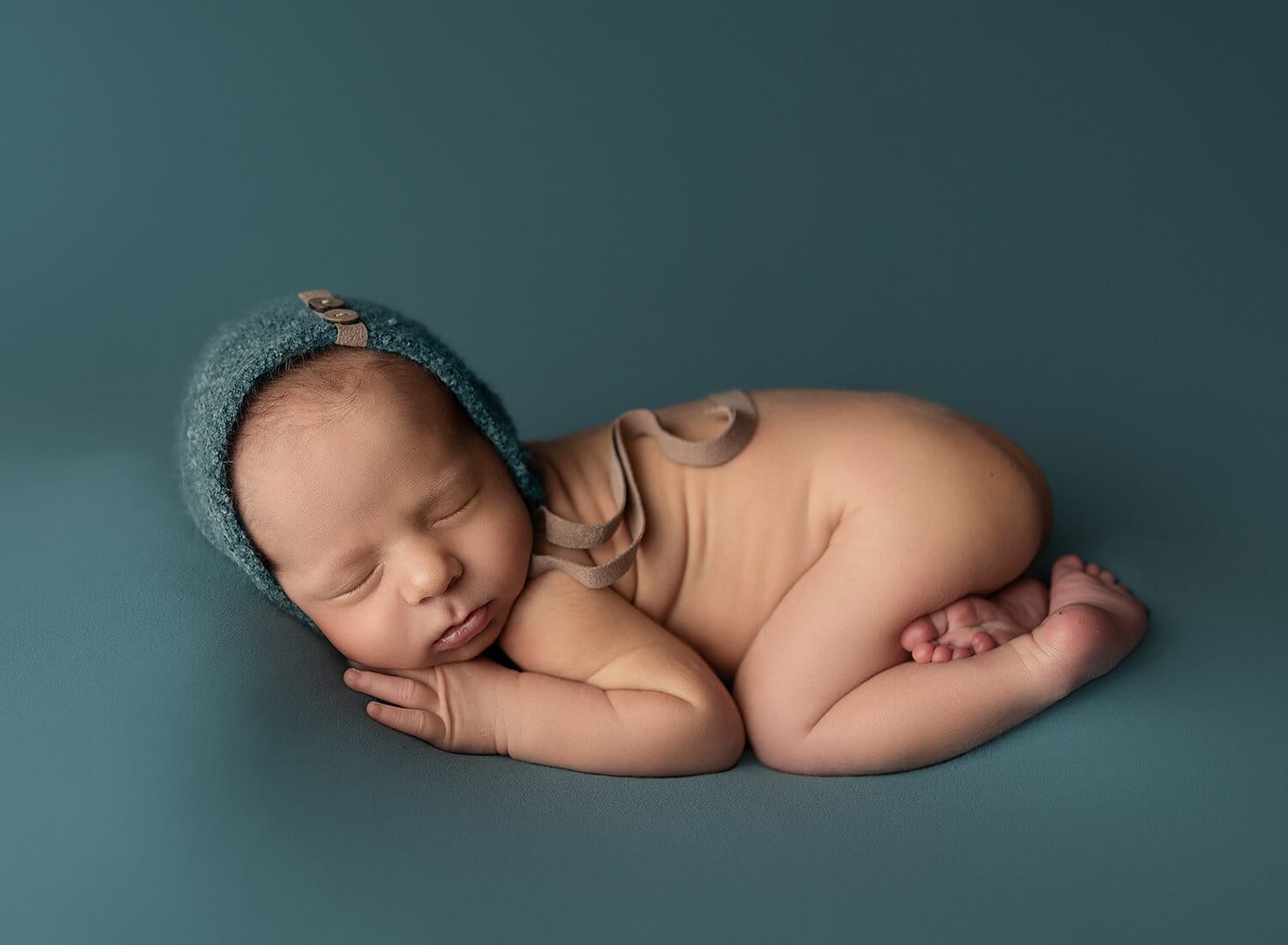 The photo shows a newborn lying on his tummy on a peacock-colored backdrop with a bonnet to match the Baton Rouge newborn photographer.