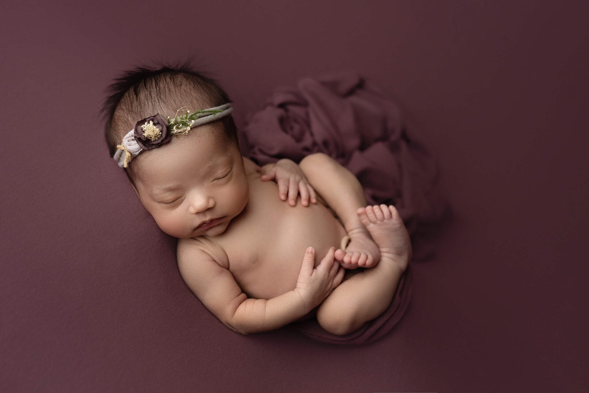 A newborn baby girl in the studio curled up in a womb pose with a floral headband.